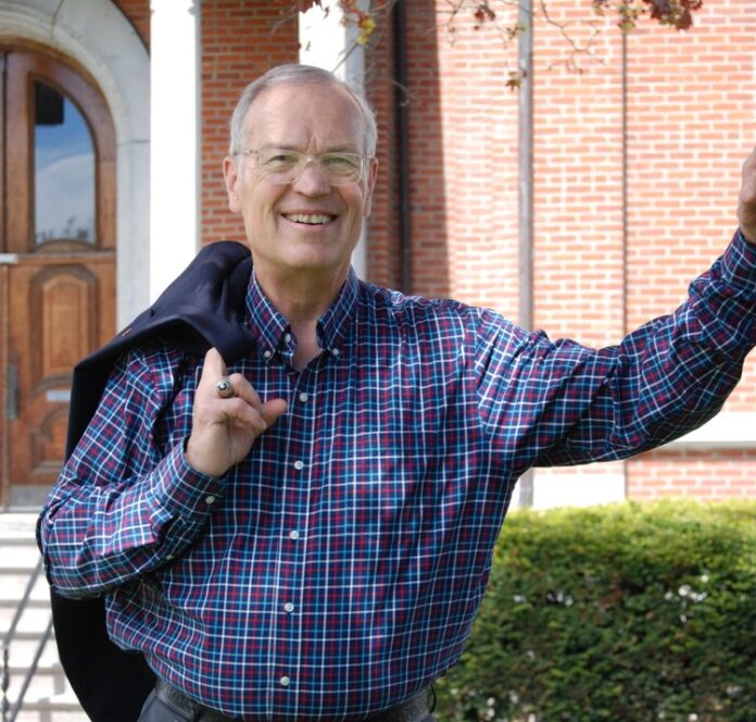 Smiling older man in a plaid shirt raises his hands outdoors in front of a brick wall.