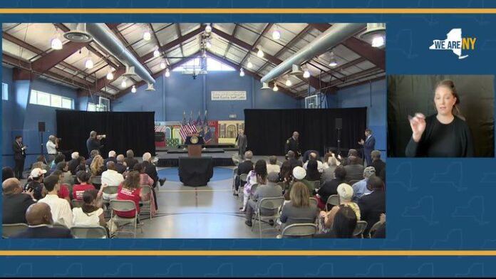 Audience seated in a hall facing a stage while a sign-language interpreter signs on a screen.