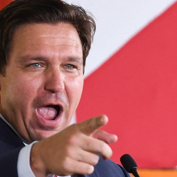Person in a suit speaking at a microphone and pointing toward the camera
