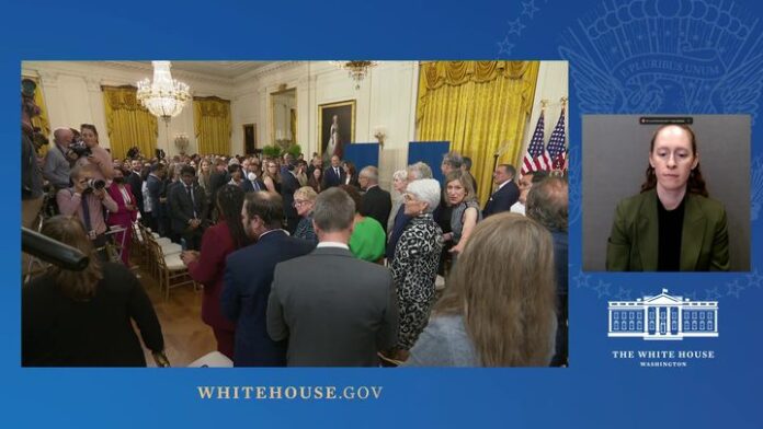 Crowd in formal White House room with officials near podium, inset video of speaker