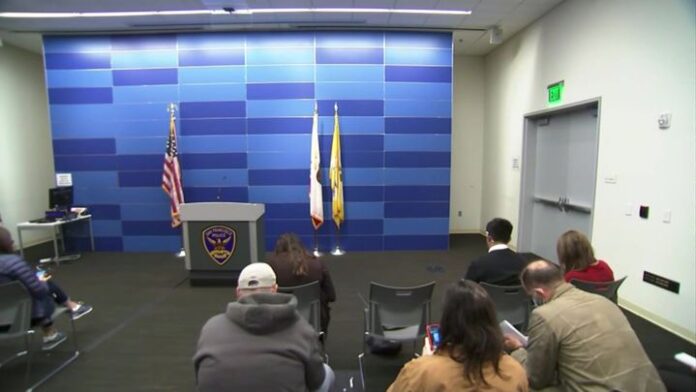 People seated facing a podium with U.S. and flag banners in a press room.