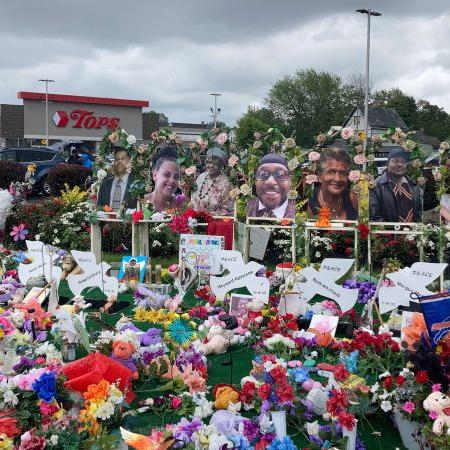 People stand behind a large memorial display with flowers, cards, and framed photos.