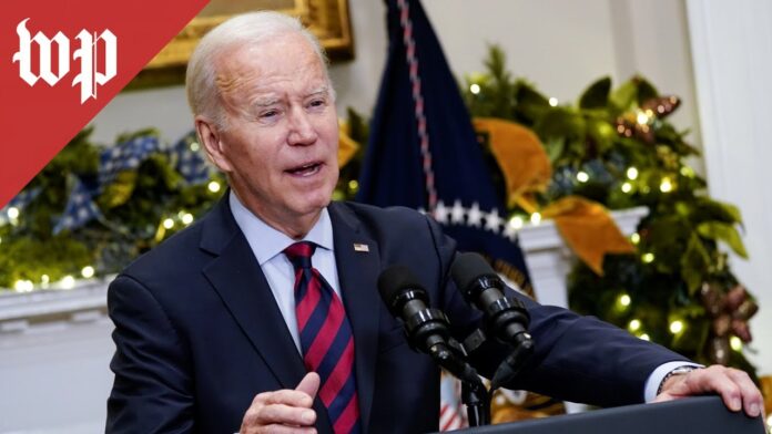 Joe Biden speaking at a podium with two microphones in front of U.S. flags and greenery.