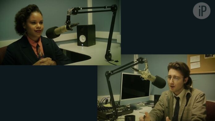 Two people sit at desks in a radio studio with boom microphones and screens.