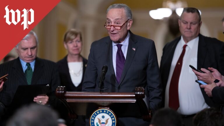 An older man in a suit speaks at a podium with microphones as people watch.