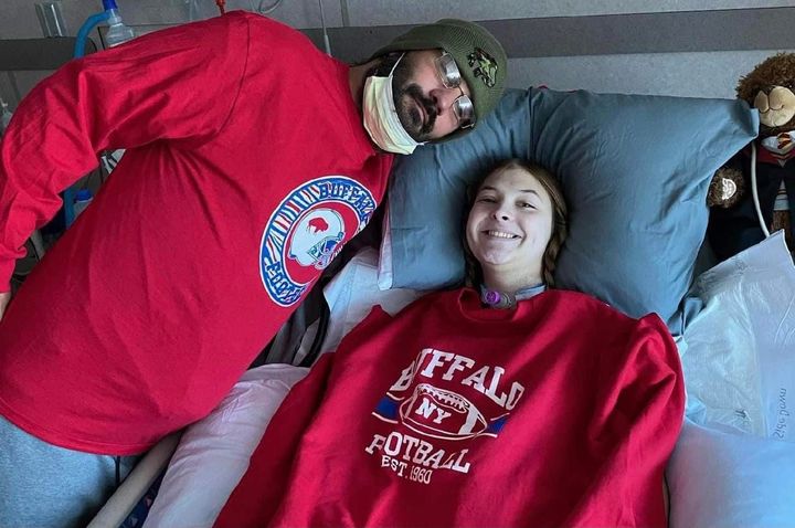 Two people in red hoodies, one wearing a cap and sunglasses, reclining on a couch.