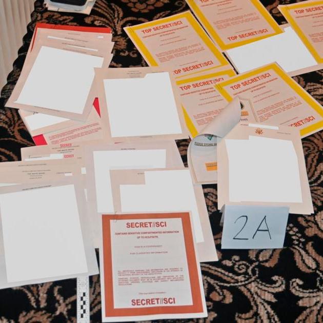 Assorted printed forms and folders labeled with “TOP SECRET/SECRET/SCI” on a patterned tablecloth.