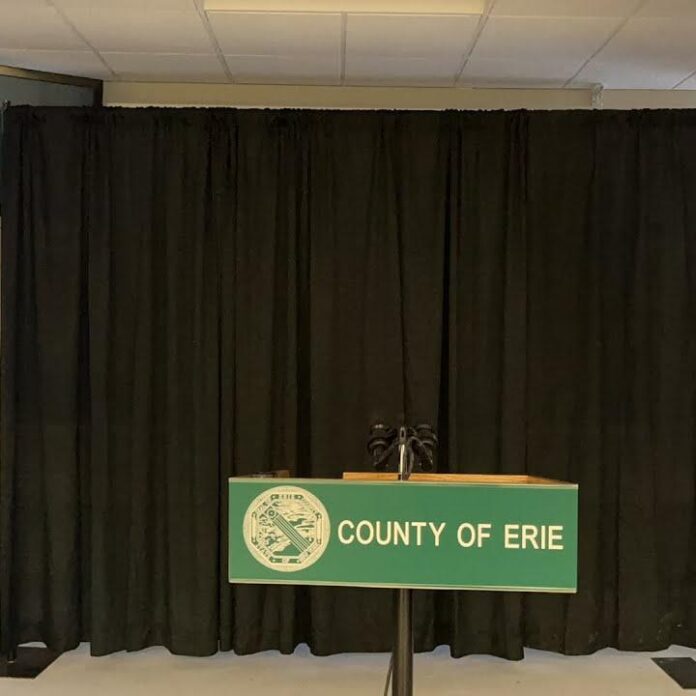 Green podium sign reading “COUNTY OF ERIE” in front of black stage curtains.