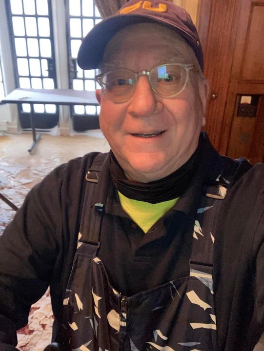 Smiling person wearing glasses, a cap, and a dark jacket indoors.