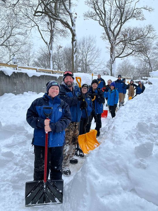 People in winter jackets pose outdoors in a snowy area with shovels