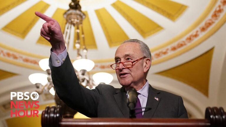 Older man in a suit gestures with his hand while speaking at a podium.