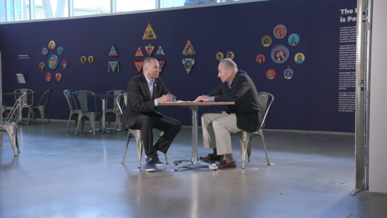 Two men in suits shake hands while seated at a table in a room with chairs and a backdrop.