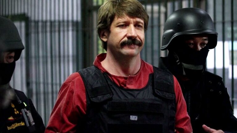 Man in red shirt and black tactical vest stands with guards wearing helmets and face coverings.