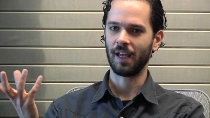 Man in dark shirt speaking and raising one hand in an interview setting