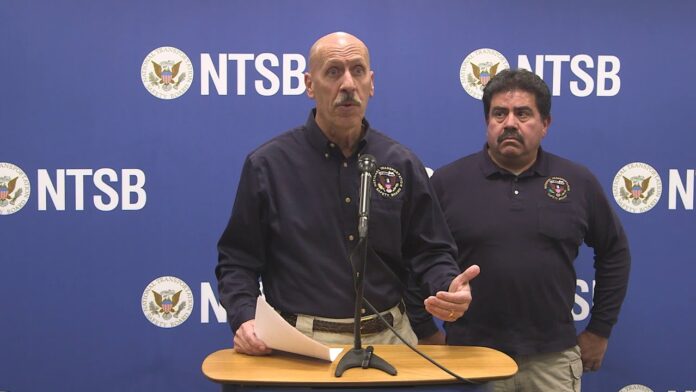 Two men stand behind a table with microphones in front of NTSB backdrop.