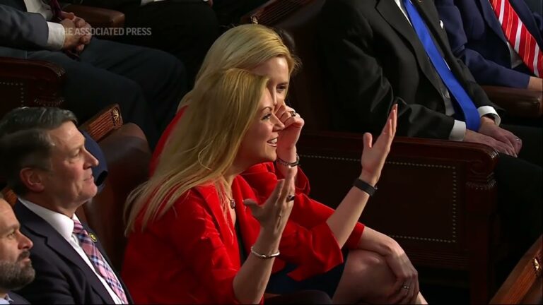 Blonde woman in a red jacket speaks and gestures in a seated chamber audience
