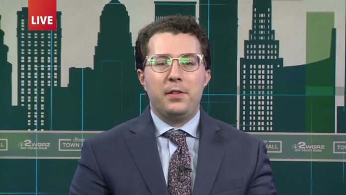 Man in a suit and glasses speaking in front of a studio city skyline graphic.