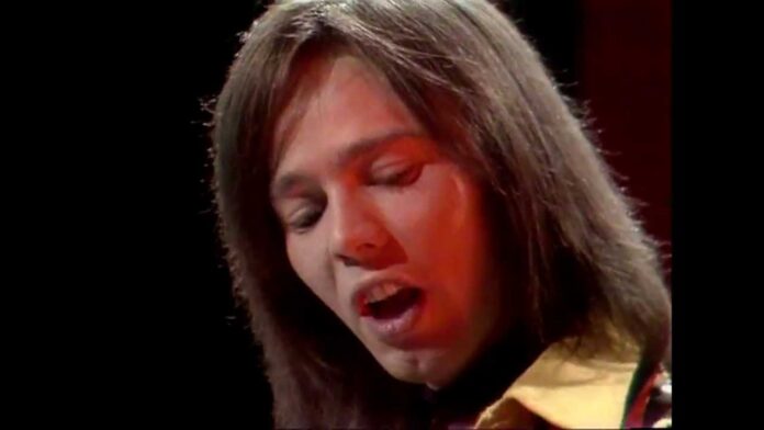Person with shoulder-length hair speaking with eyes closed under red lighting.