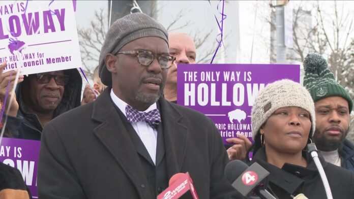 Man in suit and bow tie speaks outdoors at a rally, flanked by campaign signs and people