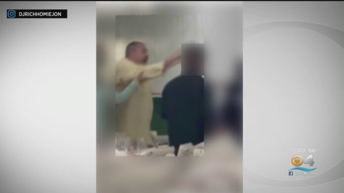 Blurred video frame shows two people near an indoor table with a tense moment.