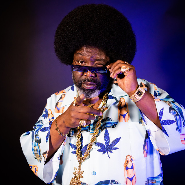 Musician in a large afro and patterned shirt holds a phone to his ear against a purple background