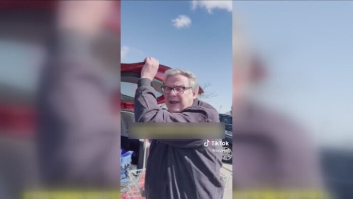 Older person waving with a raised hand outdoors near a red vehicle.