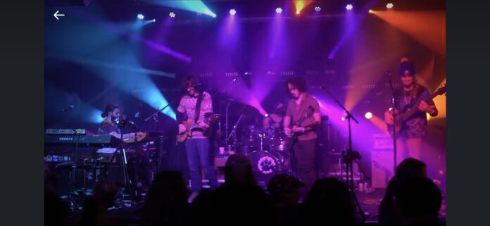 Musicians perform onstage with guitars and drums as colorful lights illuminate the crowd.