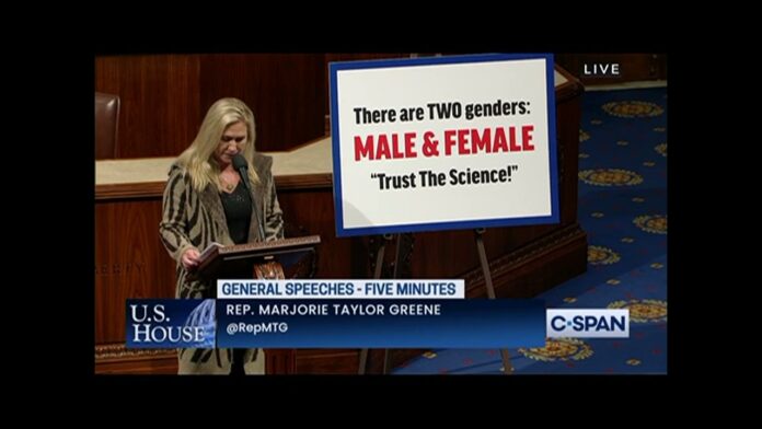 Woman speaks at a podium while a screen shows “Two genders: male & female”.