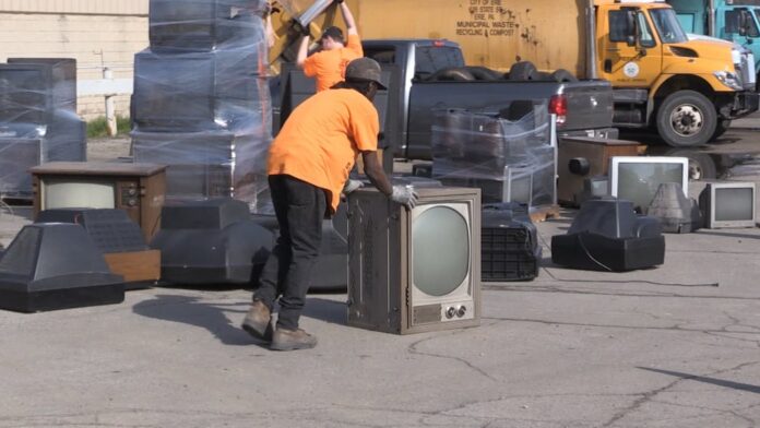 Workers wearing orange gear move a CRT television from a metal crate outdoors