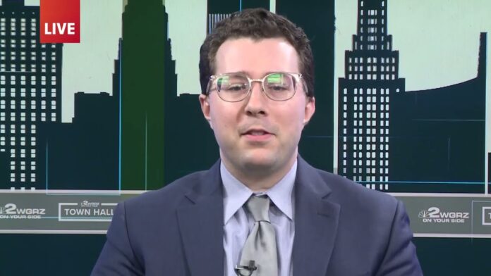 Reporter in a suit and tie speaking on camera with a city skyline backdrop.