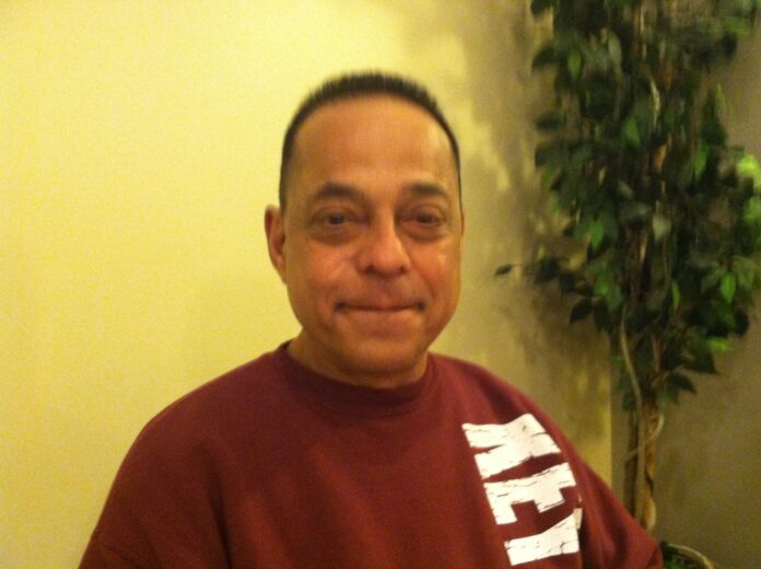 Smiling middle-aged man seated indoors wearing a maroon T-shirt with a white graphic