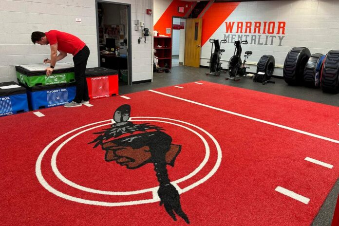 Person adjusting a black box on a red training mat inside a gym room.