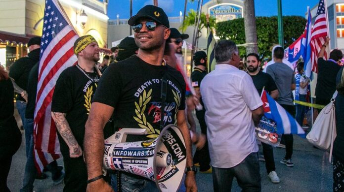 Man in sunglasses and black cap holds a microphone amid crowd with U.S. flags.