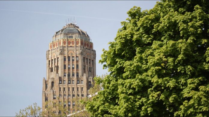 Tall historic building facade with flag-like trim, partly framed by green tree leaves.