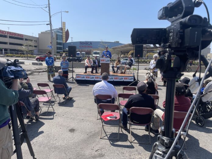 Outdoor press-style stage with speakers, microphones, seated attendees, and Seneca Casino in background