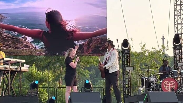 Two performers sing on an outdoor stage with instruments and large projection screens.