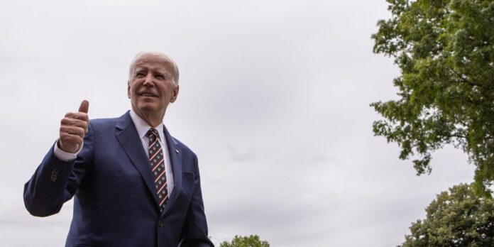 Man in a blue suit and patterned tie gives a thumbs-up outdoors under cloudy sky.