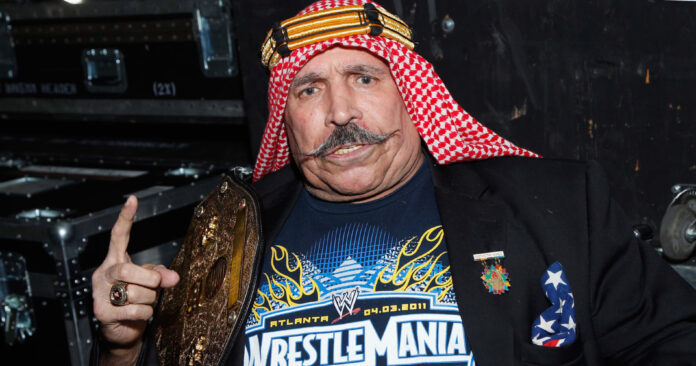 Man in keffiyeh and sports jacket holds a gold championship belt and points at camera.