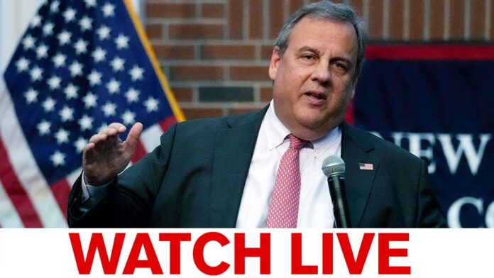 Speaker in suit at microphone with U.S. flag behind, overlaid with “WATCH LIVE”.
