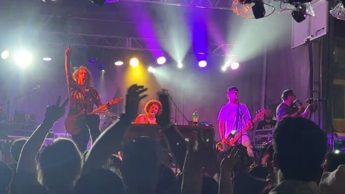 Musicians play guitars and sing on a lit concert stage with an audience in front.