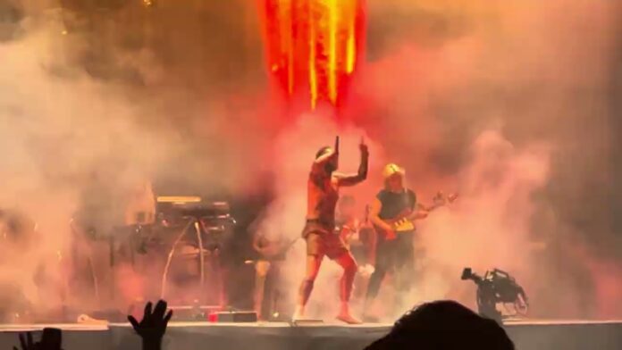 Band members on a concert stage as smoke and bright orange lights fill the scene.