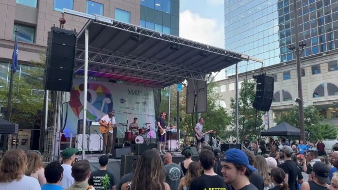 Musicians play on an outdoor stage with a crowd watching in front of downtown buildings.