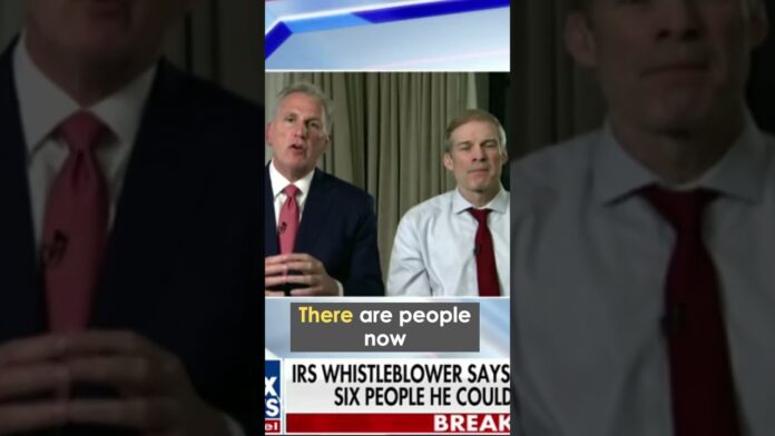 Two men in suits speak at a press event, shown within a captioned news clip.
