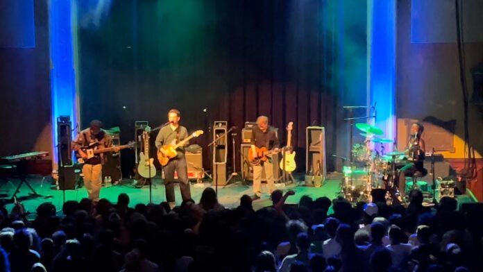 Musicians play guitars on a concert stage as an audience watches.