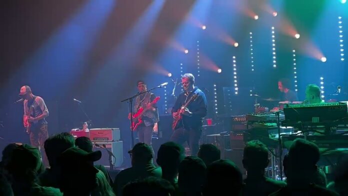 Musicians play guitars and keyboards under blue stage lights with an audience in foreground