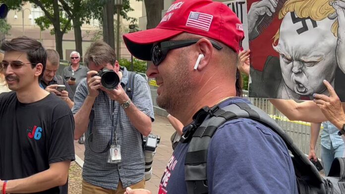 Man in red cap and sunglasses speaks while a crowd films beside a mural with a raised fist.