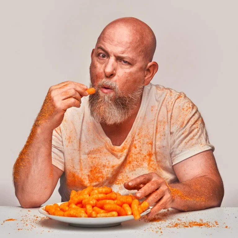 Bald man with a beard raises an orange snack above a plate covered in orange dust