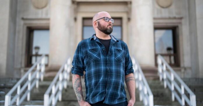 Man in plaid shirt and glasses stands on steps in front of a stone building