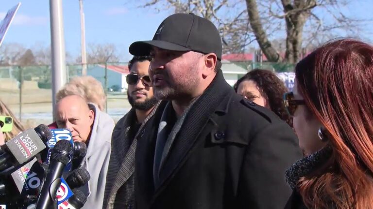 Man in a cap speaks outdoors into microphones with reporters nearby.