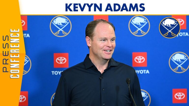 Man at a podium with microphone in front of a blue sponsor backdrop